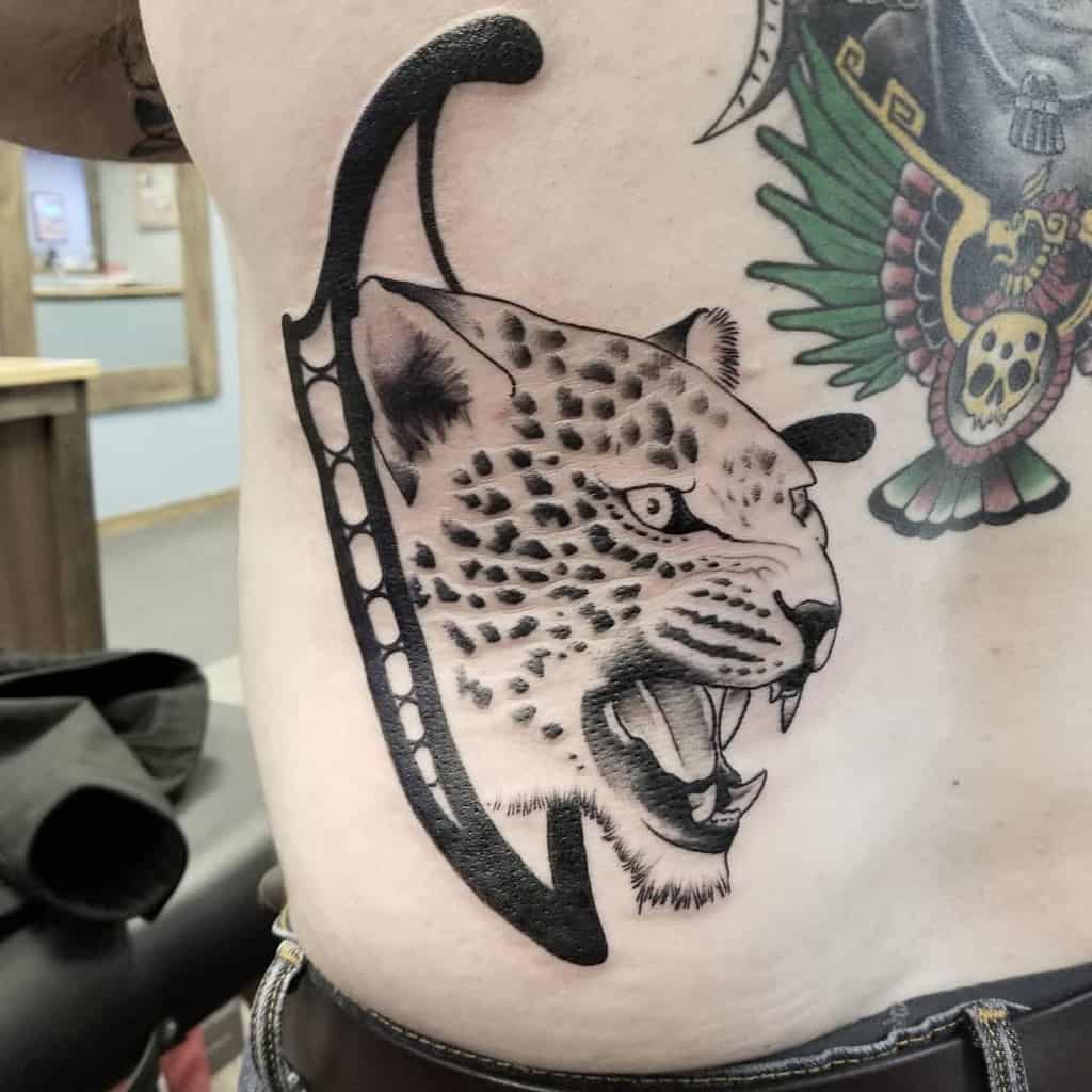 A bold black and grey tattoo of a snarling jaguar emerging from a stylized curved shape, placed on the ribs with intricate shading and detail.