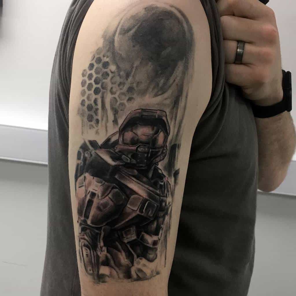 Black and gray Halo tattoo of Master Chief with a battle-worn look, featuring a planetary backdrop and hexagonal armor details on the upper arm.