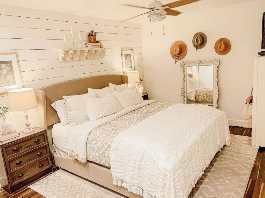 Cozy farmhouse bedroom with a large bed, wood accents, and hats hung on the wall; neutral tones and warm lighting