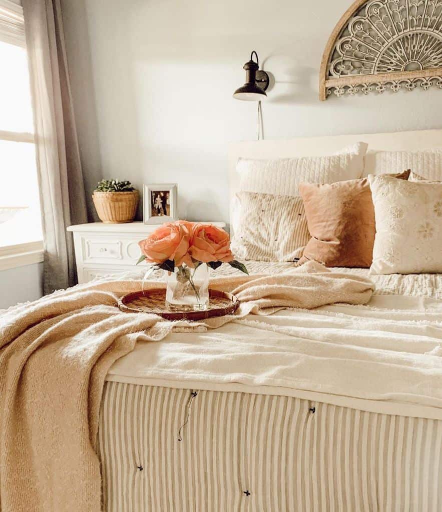 Farmhouse-style cozy bedroom with a made bed, decorative pillows, pink flowers in a vase, and a sunlit window
