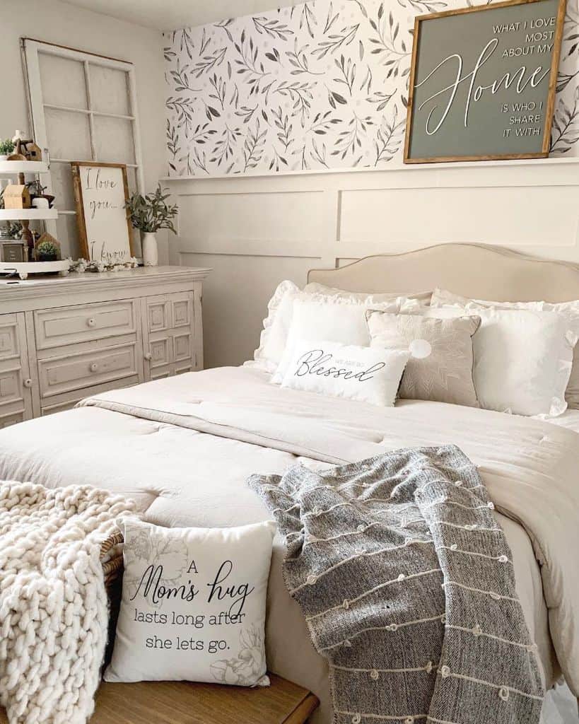 Cozy farmhouse bedroom with decorative pillows, a knitted throw, leafy wallpaper, and wall art about home and hugs
