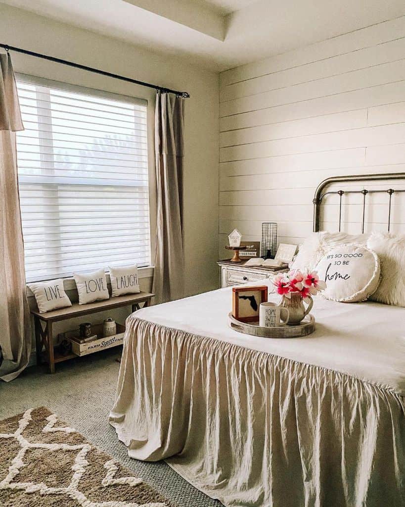 Cozy farmhouse bedroom with a metal bed, decorative pillows, and a tray; beige curtains and rug accent the room