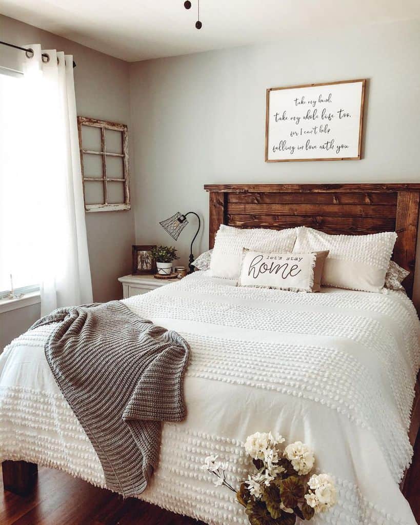 Farmhouse-style bedroom with a wooden bed, white bedding, gray blanket, and decor; sunlit window on the left
