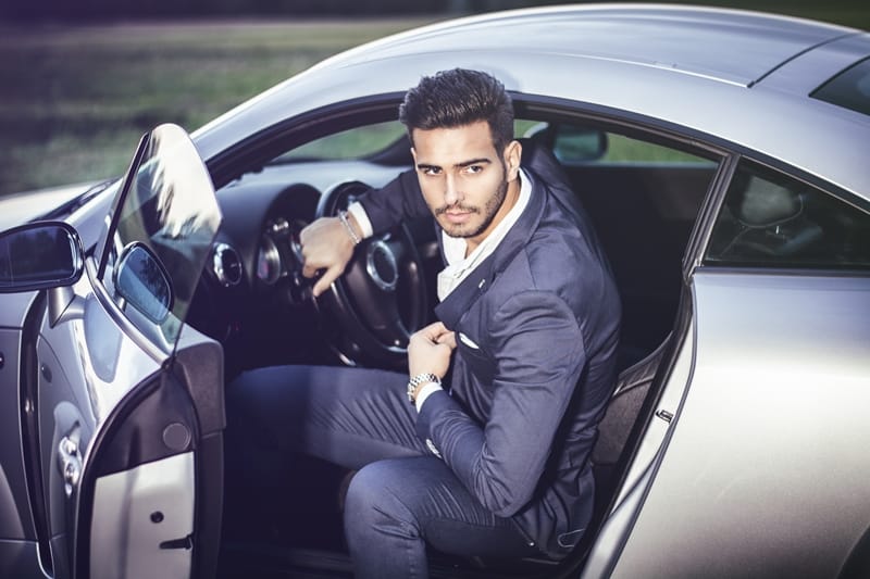 A man in a blue suit sits in the driver's seat of a silver sports car