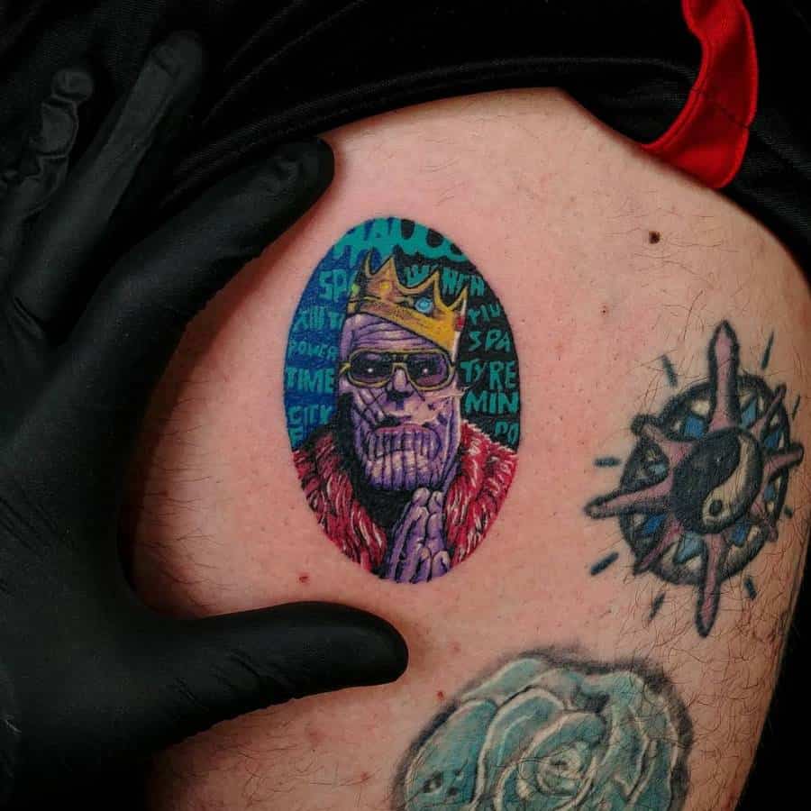 Stylized Thanos tattoo featuring a crown, sunglasses, and a fur coat, with a graffiti-style background of cosmic-themed words