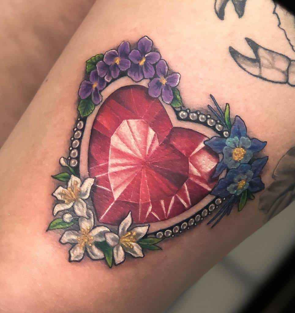 Tattoo of a red heart-shaped gem with purple, blue and white flowers, surrounded by a beaded border