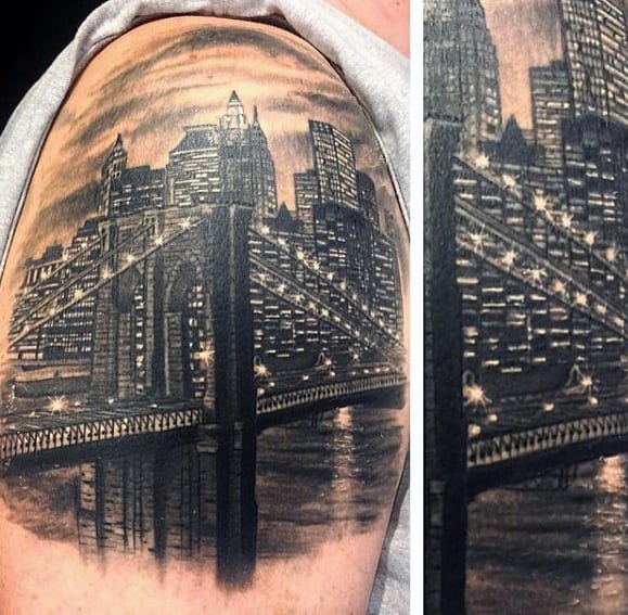Detailed upper arm tattoo of the Brooklyn Bridge at night with the New York City skyline illuminated by glowing lights and reflections.