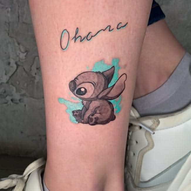 Tattoo of Stitch from Lilo & Stitch with the word "Ohana" above it on a leg