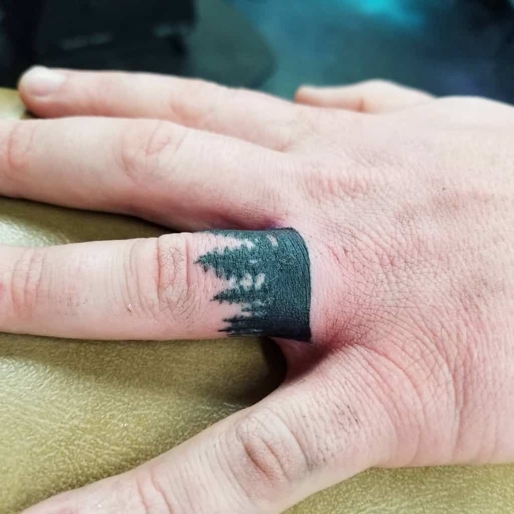 A bold, dark ring tattoo of a forest scene with tree silhouettes, in a solid band of black ink