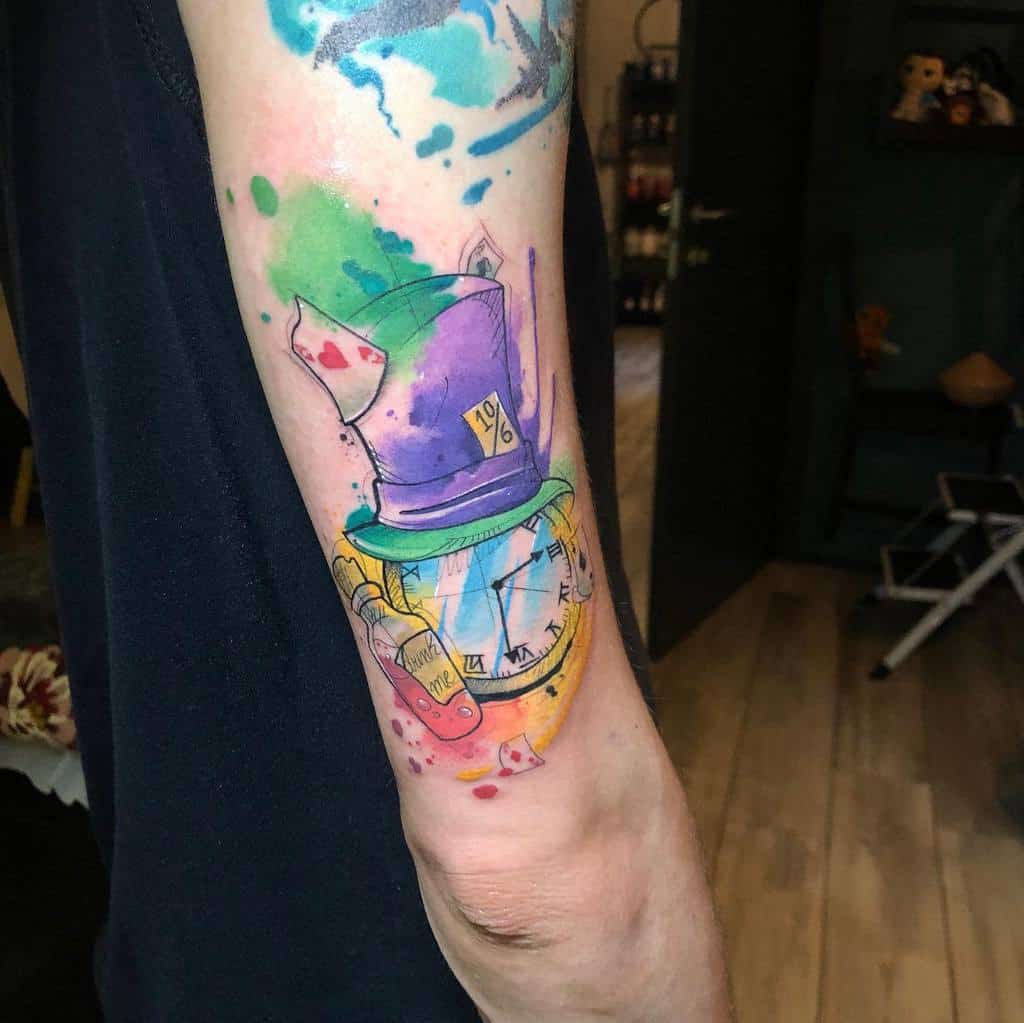 A colorful tattoo featuring the Mad Hatter's hat, a pocket watch, and a "Drink Me" bottle, playing cards with vibrant watercolor splashes