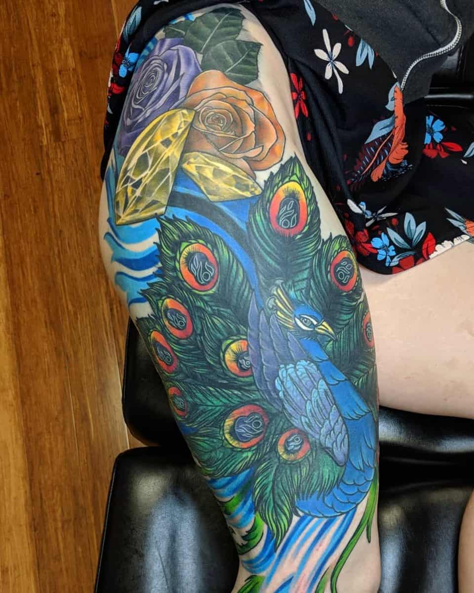 Colorful tattoo featuring a blue peacock, rose, and crystals on a thigh