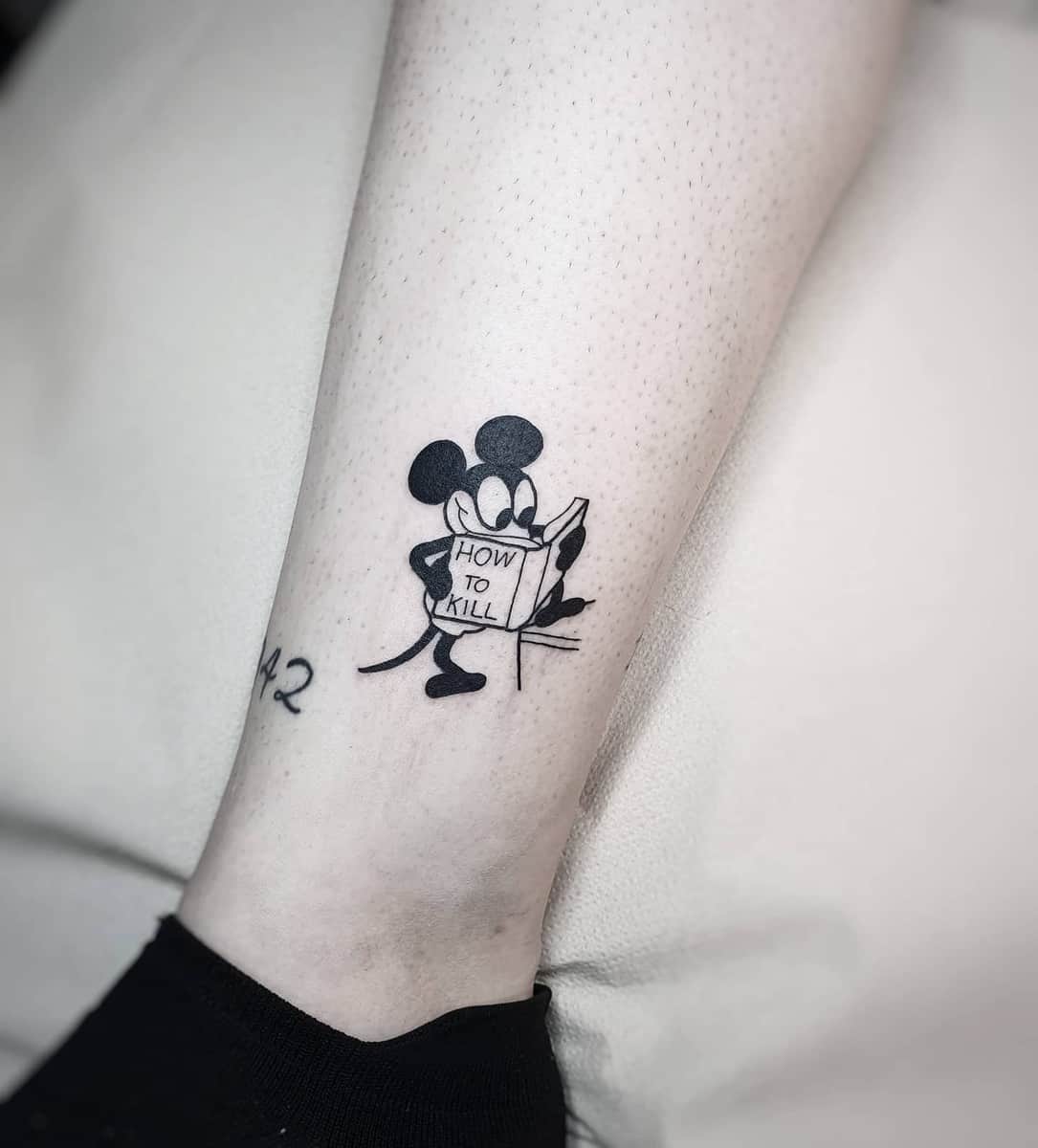 Tattoo of a mouse reading a book titled "How to Kill" on a leg