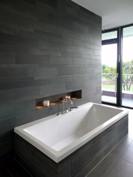 Built-in bathtub with dark grey matte porcelain tile wall and recessed shelf.