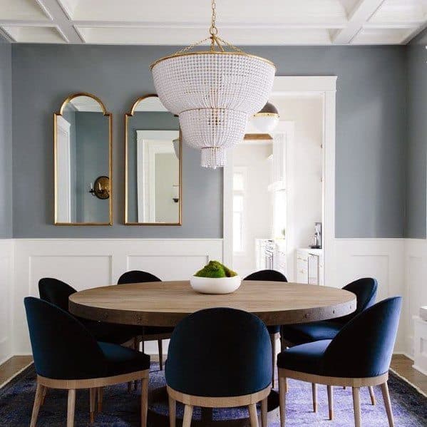 Elegant dining room with a round wooden table, chair rail, blue chairs, crystal chandelier, and mirrors on gray walls