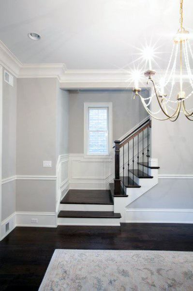 Elegant staircase with dark wood steps, white trim, and a chandelier; gray walls with chair rail and a window in the background