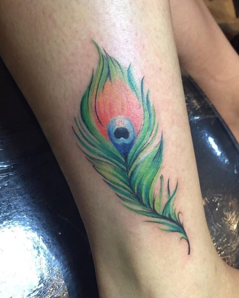 Vibrant peacock feather tattoo on the lower leg, featuring green, blue, and orange hues with soft shading and delicate linework.