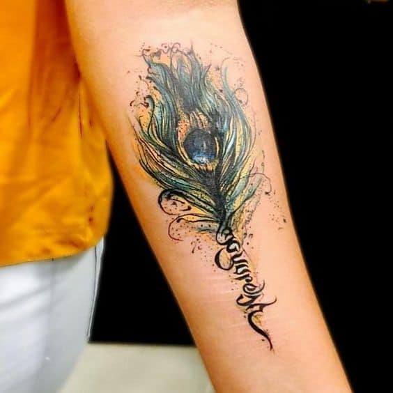Artistic peacock feather tattoo on the forearm with watercolor-style splashes, intricate detailing, and the word 'freedom' elegantly scripted.