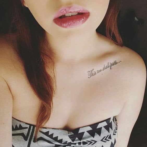 A "This Too Shall Pass" tattoo in cursive on the collarbone area, with ellipses at the end.