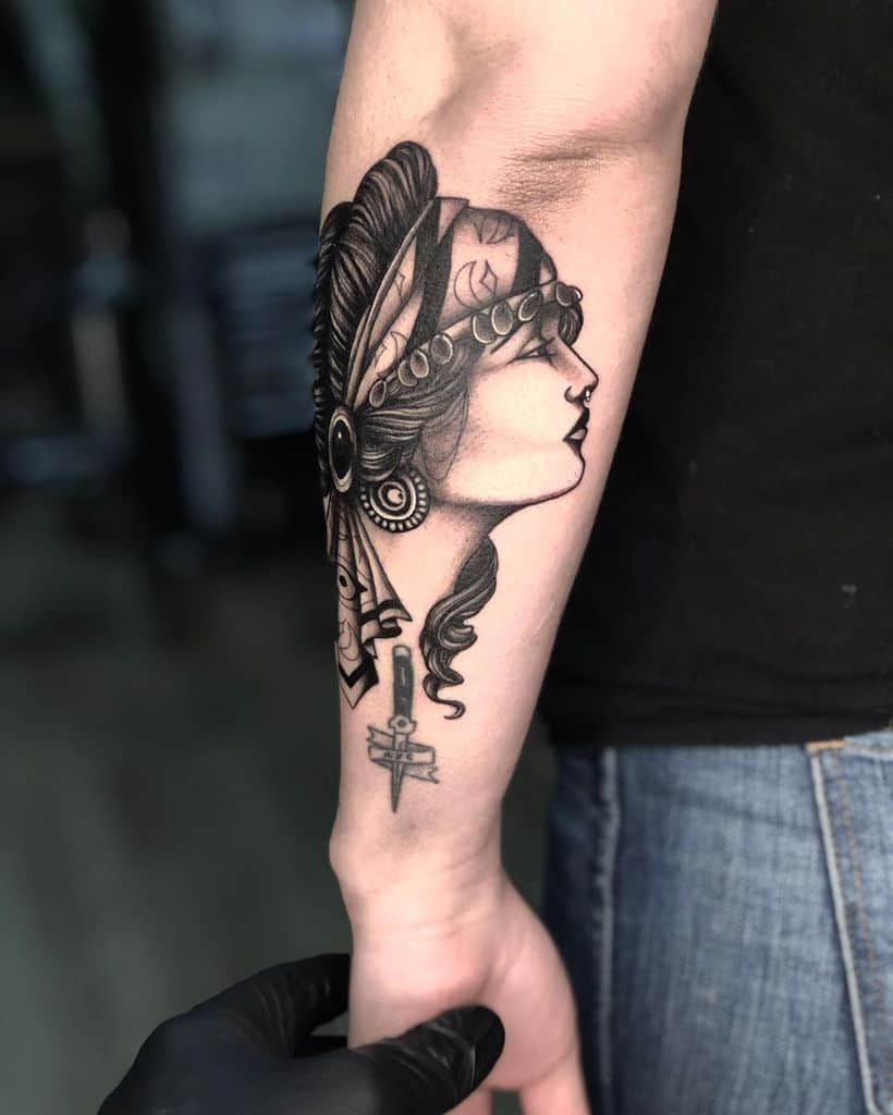Black and grey tattoo of a gypsy woman in profile with a beaded headscarf, detailed shading, and a small dagger design on the forearm.
