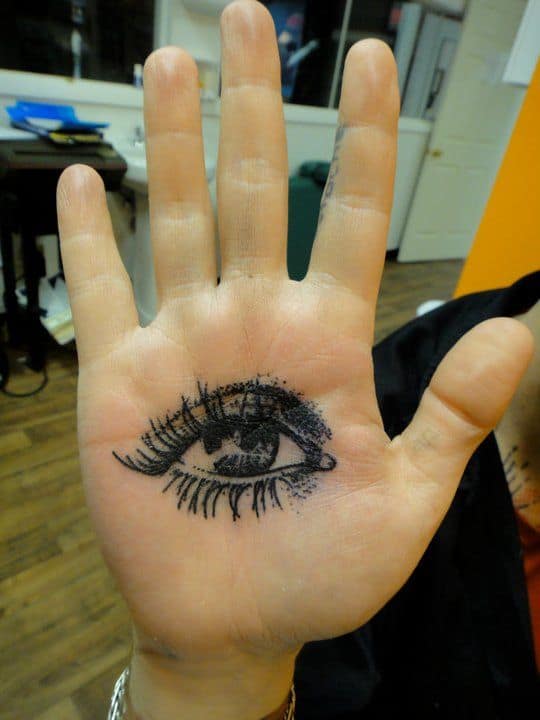 Tattoo of a realistic eye inked in black on the palm, with detailed eyelashes and shading.