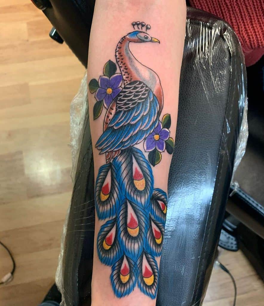 Peacock tattoo with vibrant blue feathers, red accents, and purple flowers on the forearm.