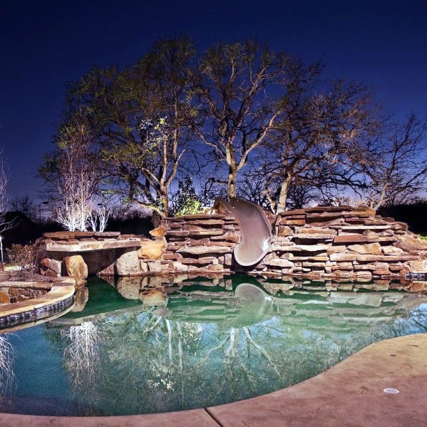 Backyard pool with rock waterfall and slide, surrounded by trees, under a twilight sky
