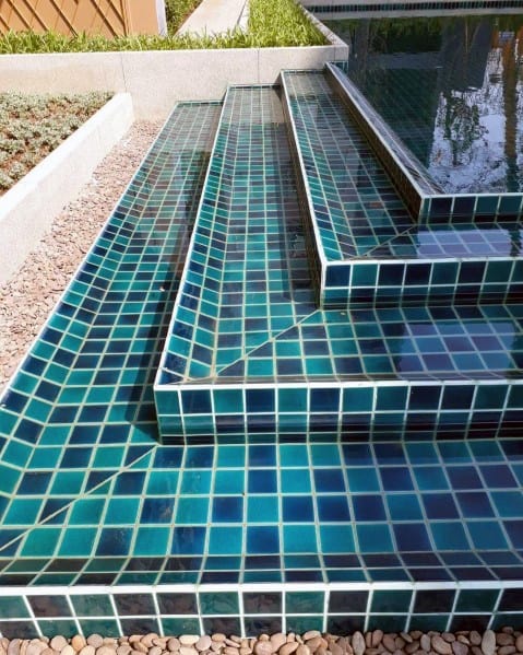 Green and blue mosaic-tiled pool steps with sleek design and surrounding pebble landscaping.