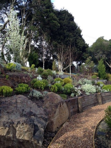 Sloped garden with natural boulders, wooden retaining walls, gravel pathways, and lush plants creating a rustic and serene landscape