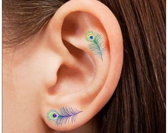 Two small peacock feather tattoos inside and below the ear, featuring delicate green, blue, and purple hues with fine detailing for a subtle look.