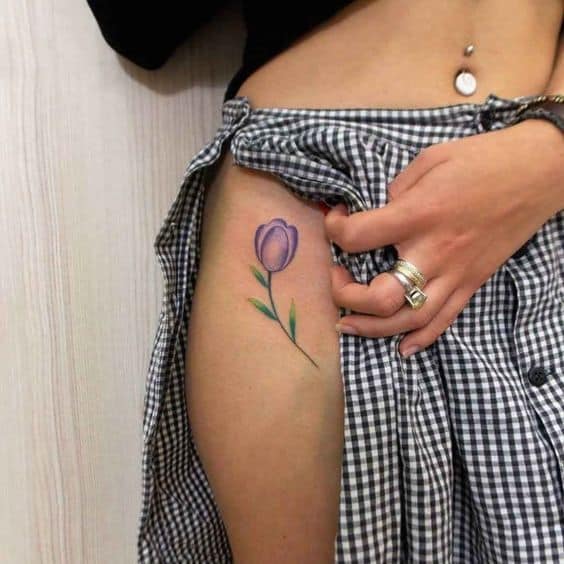 A delicate purple tulip tattoo on the upper thigh, featuring fine shading, a thin green stem, and small leaves for a soft and elegant floral design.