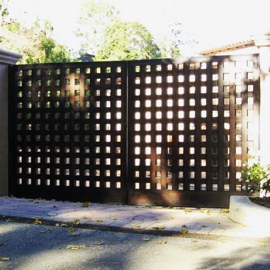 A wooden lattice gate casts shadows onto the ground, surrounded by trees and a brick pathway