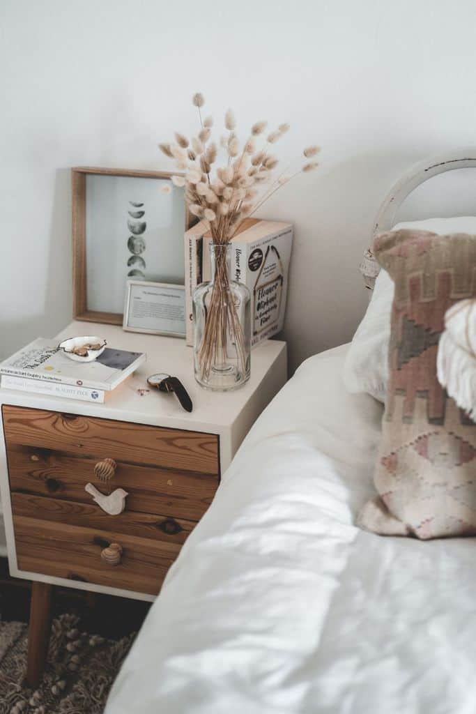 Rustic nightstand with dried flowers, books, and cozy decor beside a white bed.