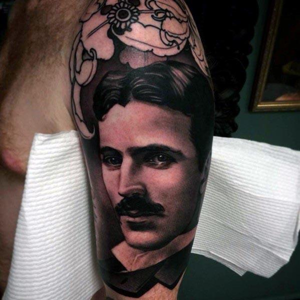 Black and grey tattoo of Nikola Tesla with realistic facial detail, framed by abstract mechanical elements and soft shading above.