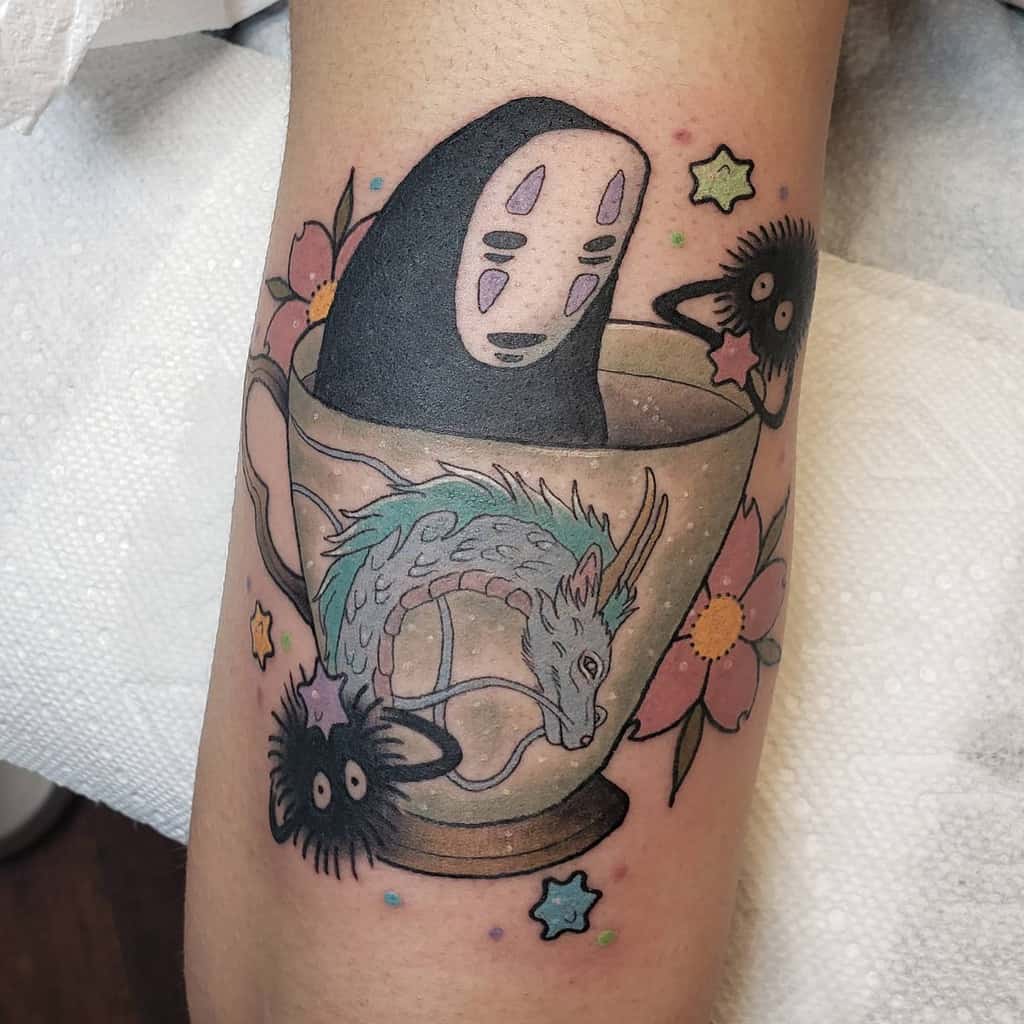 Whimsical Spirited Away tattoo featuring No-Face in a teacup, Haku in dragon form, soot sprites, star candies, and floral accents