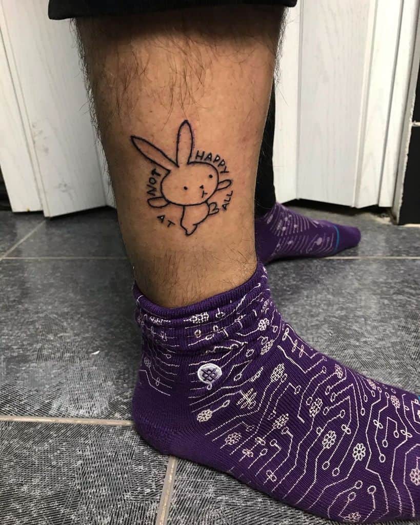 A simple black ink tattoo of a sad bunny with the phrase 'Not Happy At All' written around it, placed on the lower leg.
