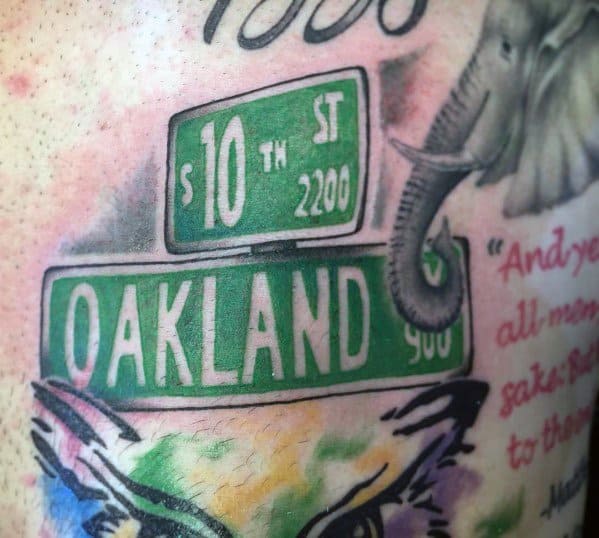 Colorful tattoo featuring green 'Oakland' and '10th St' street signs, with an elephant trunk and artistic watercolor-style elements.