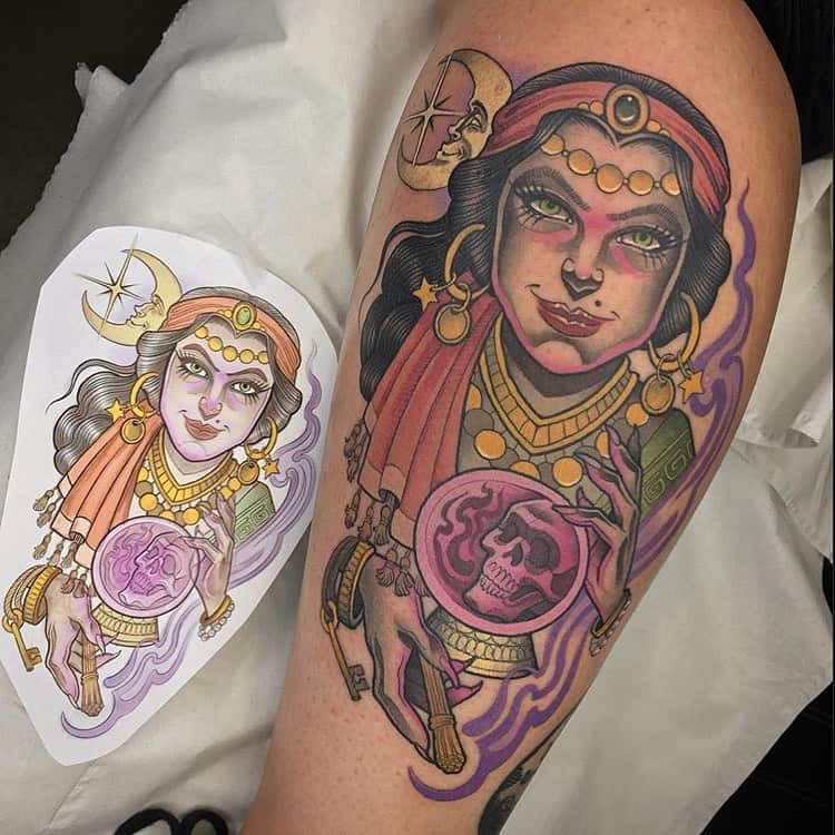 Colorful tattoo of a mystical fortune teller with glowing green eyes, gold jewelry, and a crystal ball revealing a skull, placed on the arm.
