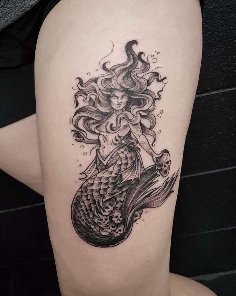 Black and grey tattoo of a fierce mermaid with wild flowing hair, skull-adorned tail, and a menacing expression, surrounded by bubbles.