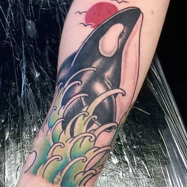 Vibrant tattoo on the forearm, featuring an orca whale emerging from colorful waves beneath a red sun, blending dynamic movement with a serene atmosphere.