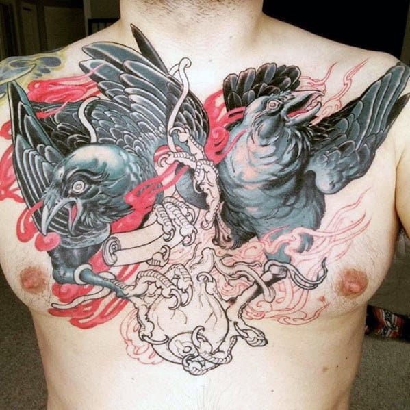Vivid chest tattoo of Huginn and Muninn emerging from a detailed anatomical heart, with bold black feathers and swirling red accents.