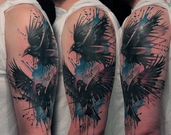 Watercolor-style upper arm tattoo of Huginn and Muninn in flight, with bold black strokes, blue splashes, and abstract paint splatters.