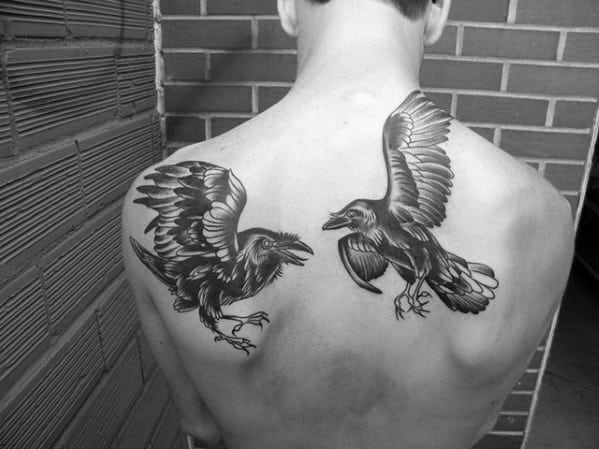 Black and grey back tattoo of Huginn and Muninn in flight, positioned on each shoulder blade with detailed feathers and fierce expressions.