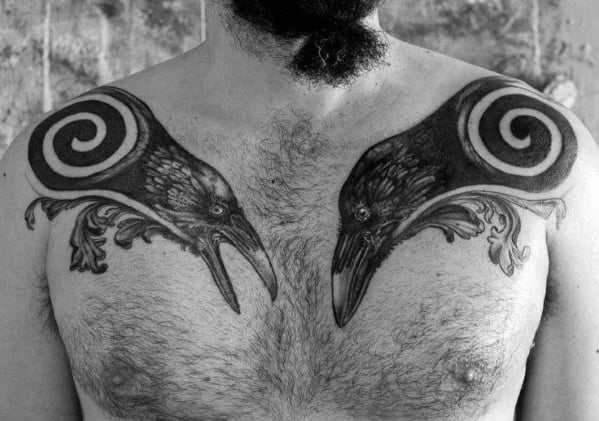 Stylized chest tattoo of Huginn and Muninn facing each other, with spiral patterns above their heads and ornamental detailing below.