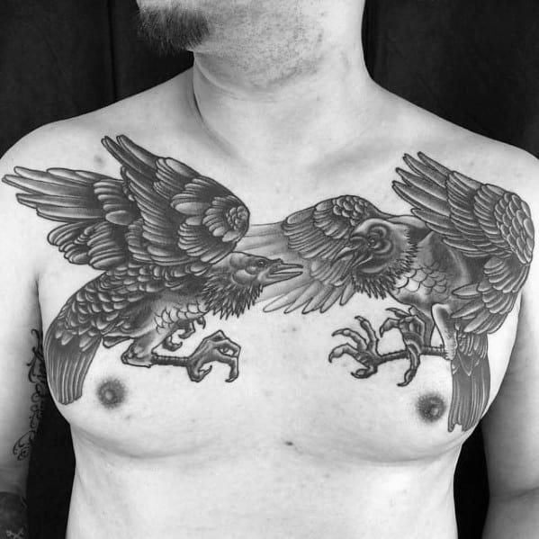 Black and grey chest tattoo of Huginn and Muninn mid-flight, facing each other with outstretched wings and detailed feathers and claws.