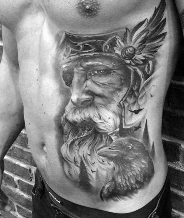 Realistic black and grey side torso tattoo of Odin with a winged helmet, intense gaze, and a raven beneath, symbolizing wisdom and watchfulness.