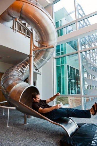 Person sliding down a spiral slide indoors, cup in hand, against large windows and sleek modern architecture