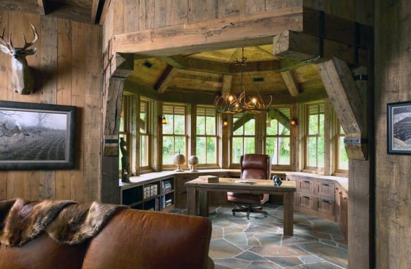 Rustic office with wooden beams, large windows, leather chair, and antler chandelier, cozy natural lighting