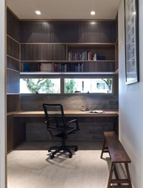 Minimalist home office with wooden shelves, a built-in desk, a black chair, and bench overlooking greenery through a window