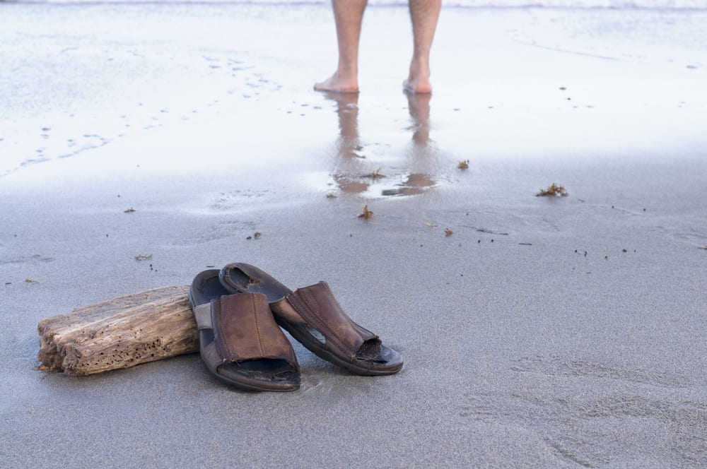 A pair of men's brown slide sandals propped up on driftwood on the beach