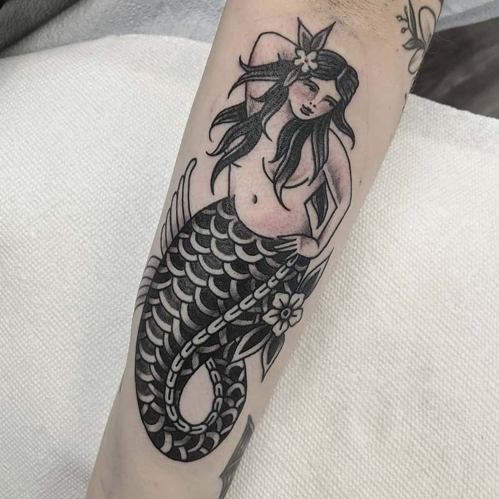 Black and grey traditional tattoo of a mermaid with flowing hair, a flower in her hair, and a curved tail decorated with floral details.